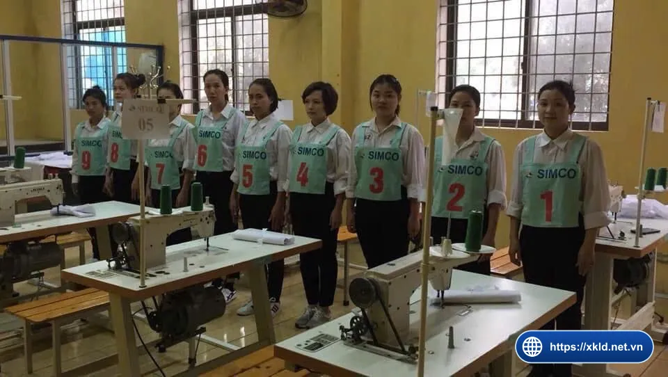 Chín lao động nữ mặc áo sơ mi trắng, đeo yếm xanh ghi SIMCO Sông Đà số 1 đến 9, đứng thành hàng cạnh các bàn máy may công nghiệp trong phòng học có cửa sổ, trong buổi thi tuyển đơn hàng may mặc của SIMCO Sông Đà