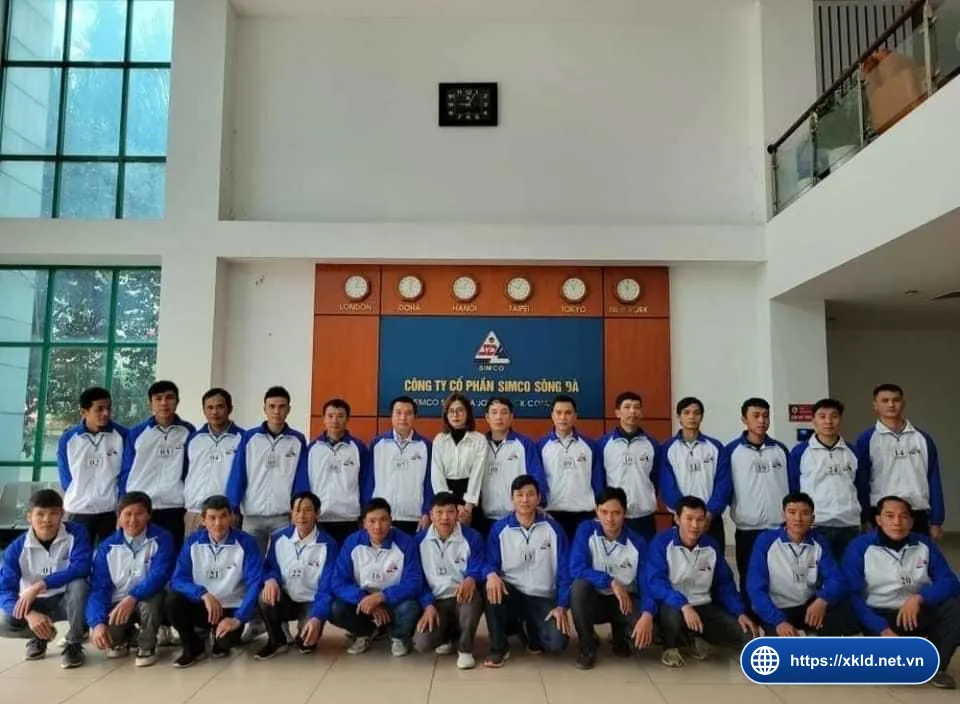 Hơn hai mươi lao động nam mặc áo khoác xanh trắng, đeo số báo danh, xếp hai hàng đứng và ngồi xổm cùng một nhân viên nữ trước bảng hiệu SIMCO Sông Đà trong sảnh văn phòng