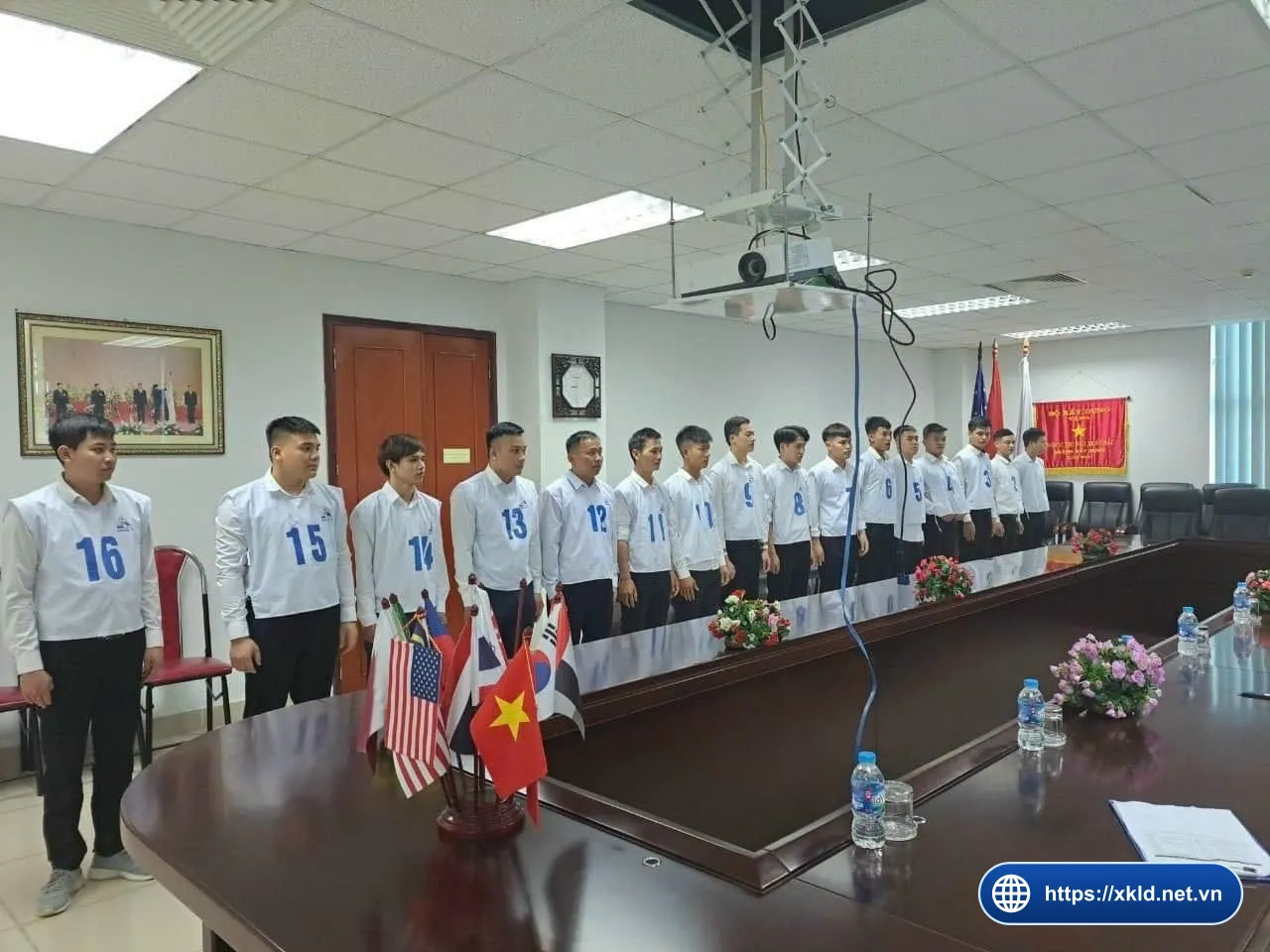 Nhóm lao động nam mặc áo sơ mi trắng, đeo yếm số từ 3 đến 16, đứng thành hàng trong phòng họp có bàn dài, cờ nhiều nước đặt trên bàn, chờ đến lượt thi tuyển đơn hàng của SIMCO Sông Đà