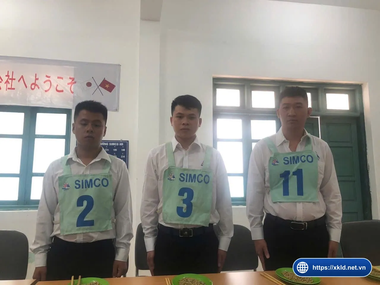 Ba lao động nam mặc áo sơ mi trắng, đeo yếm xanh ghi SIMCO Sông Đà số 2, 3 và 11, đứng sau bàn có đĩa xanh và đũa, phía sau là băng rôn tiếng Nhật với cờ Nhật Bản và cờ Việt Nam, trong buổi thi tuyển đơn hàng của SIMCO Sông Đà